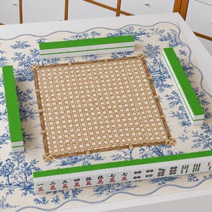May include: A mahjong set featuring green-backed tiles with white faces, some displaying red and black Chinese characters and circular patterns. The tiles are arranged on a square mat with a blue and white toile de Jouy pattern and a central woven cane-style square.