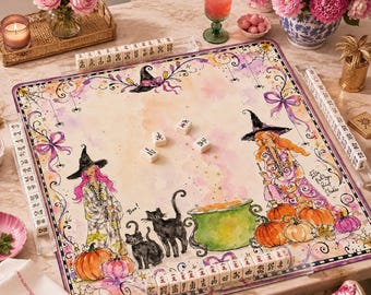May include: A square mahjong mat featuring a watercolor illustration of two witches, black cats, pumpkins, and a green cauldron. The mat is bordered by acrylic tile racks holding white mahjong tiles with black characters and symbols.