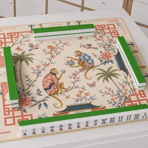 May include: A square mahjong mat featuring a cream-coloured surface with a central illustration of two monkeys in patterned clothing sitting on flowering branches. The mat includes green border accents and four rows of white and green mahjong tiles.