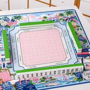 May include: A Mahjong mat featuring a coastal theme with sailboats, hydrangeas, and houses. The centre has a pink grid with text including Charleston, Play Right, and Walls Left. Green tiles are arranged on the mat, with some displaying Chinese characters.