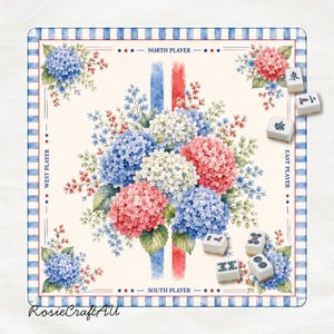 Puede incluir: Un tapete de mahjong cuadrado con un ramo central de hortensias azules, rosas y blancas en acuarela. El borde tiene rayas azules y blancas con etiquetas para los jugadores Norte, Sur, Este y Oeste. Varias fichas de mahjong blancas est&aacute;n dispersas.