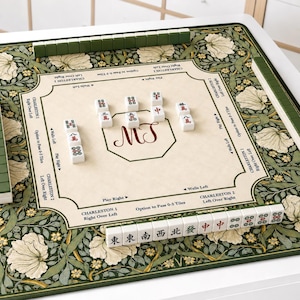 May include: A square mahjong mat featuring a cream-coloured centre with printed game instructions and the letters MT, surrounded by a dark green border with a white floral pattern. Several white mahjong tiles are arranged on the mat surface.