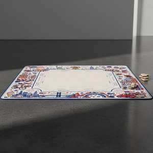 Puede incluir: Una alfombrilla de juego rectangular con un centro color crema y un borde con ilustraciones en rojo, blanco y azul de monumentos estadounidenses, una bicicleta, un cami&oacute;n y fuegos artificiales. Dos dados blancos y fichas de madera al lado.