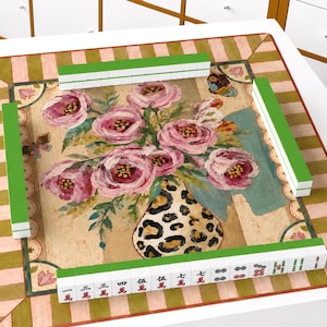 以下が含まれることがあります： 中央にヒョウ柄の花瓶に入ったピンクのピオニーの絵が描かれ、くすんだピンクとグリーンのストライプの縁取りが施された正方形の麻雀マット。マットの上には、裏面が緑色の白い麻雀牌が4列に並べられています。