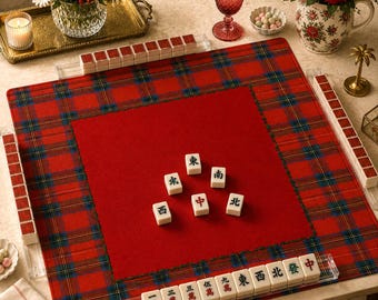 May include: A square mahjong mat featuring a red and blue plaid border with a solid red center. The mat includes clear acrylic tile racks along each side and white mahjong tiles with black and red Chinese characters arranged on the surface.