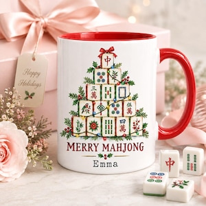 Op de afbeelding: Een witte keramische mok met een rode binnenkant en handvat, voorzien van een afbeelding van mahjongtegels gestapeld in de vorm van een kerstboom. De tekst MERRY MAHJONG en de naam Emma staan onder de boom, versierd met hulst en lichtjes.