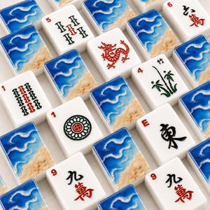 Könnte beinhalten: Eine Sammlung weißer Mahjong-Spielsteine, die in einem Raster angeordnet sind. Einige Steine zeigen blaue Meereswellen und Sandstrände, während andere rote, schwarze und grüne Symbole, Zahlen, einen Drachen sowie Bambusmotive aufweisen.