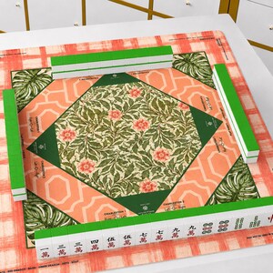 May include: A square mahjong mat featuring a peach and white plaid border, a central green diamond with a floral pattern, and four green corner sections with monstera leaves. Four rows of green and white mahjong tiles are arranged on the mat.