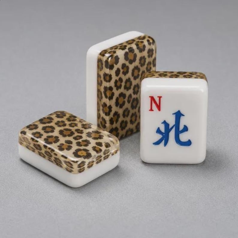 May include: Three rectangular mahjong tiles with a white base and a brown and black leopard print pattern on the back. One tile is positioned upright, showing the front face with a blue Chinese character and a red letter N printed on the white surface.