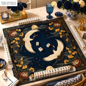 May include: A square mahjong mat featuring a dark blue background with two white cranes, golden ginkgo leaves, and floral patterns. The mat includes four clear acrylic tile racks filled with white mahjong tiles and three loose tiles in the centre.