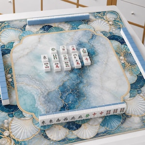 May include: A square mahjong game board featuring a blue and white marble-patterned surface with decorative seashell and pearl illustrations. The board includes four rows of light blue tiles arranged along the edges and a central cluster of tiles.