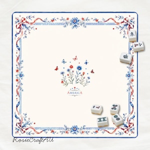 Puede incluir: Un tapete de mahjong cuadrado de color blanco roto con un borde de cintas, flores y mariposas rojas, blancas y azules. El centro muestra un arreglo floral con mariposas y la palabra AMERICA. Cinco fichas de mahjong est&aacute;n esparcidas.
