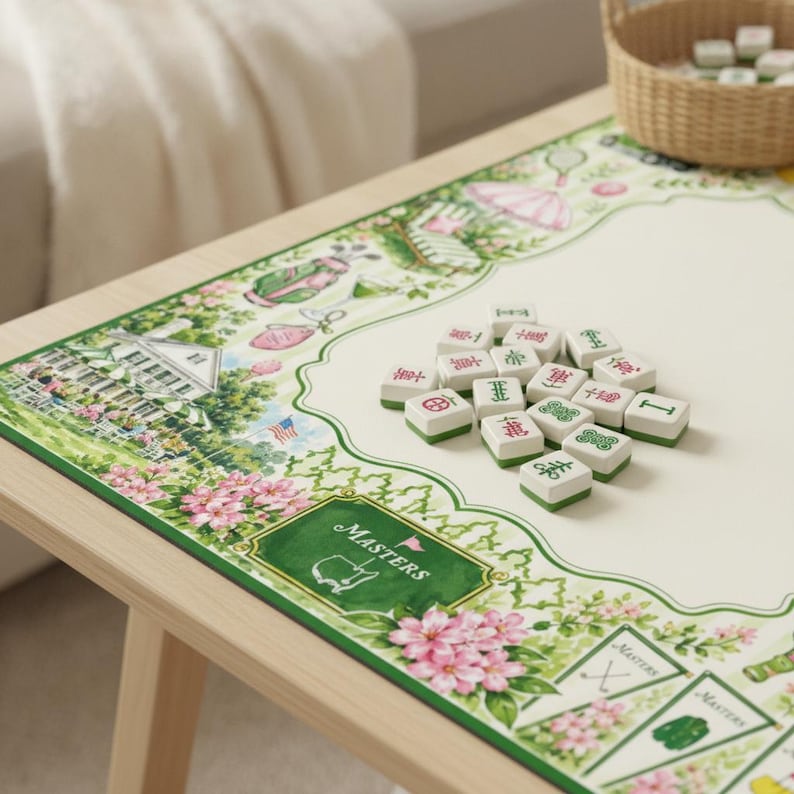 May include: A rectangular game mat featuring a cream-colored center and a decorative border with illustrations of a clubhouse, golf bags, and pink flowers. The word MASTERS is printed on a green rectangular emblem. A pile of white mahjong tiles sits on top.