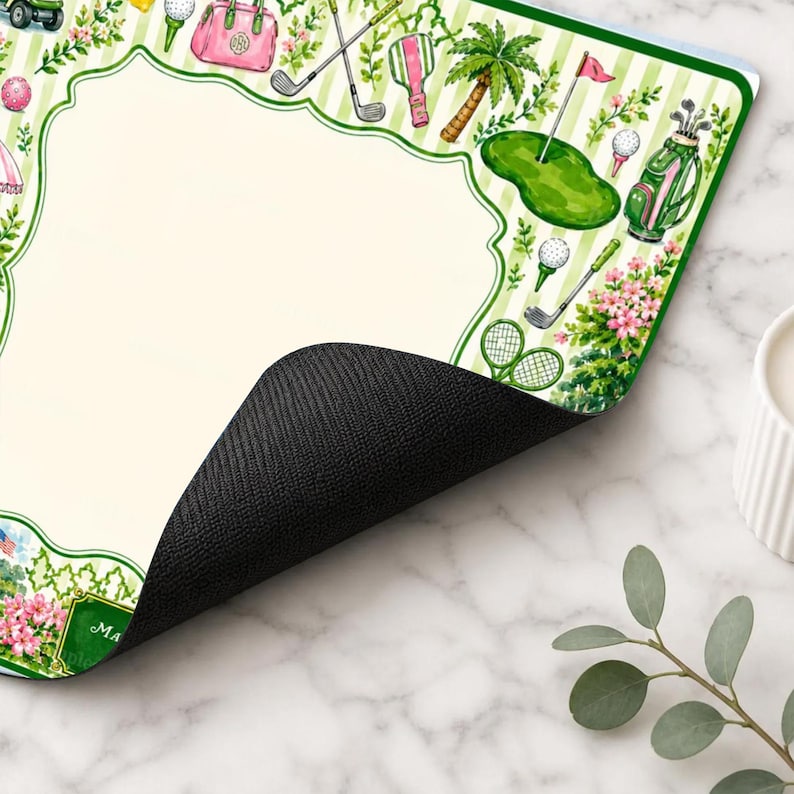 May include: A rectangular desk mat with a cream-colored center and a green and white striped border featuring watercolor illustrations of golf clubs, golf bags, golf balls, palm trees, pink flowers, and a golf green with a flag.