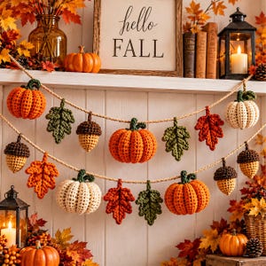 Op de afbeelding: Een tweelaagse slinger met gehaakte oranje en crèmekleurige pompoenen, bruine en beige eikels, en groene en rode herfstbladeren aan een dik touw van jute. De decoratieve slinger toont diverse herfstvormen gemaakt van garen.