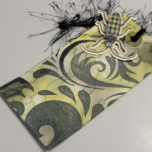 May include: A handmade gift tag with a yellow and green watercolor background and a black swirl design. A paper spider is attached to the top of the tag, and black string is attached to the top. The tag is perfect for Halloween or other spooky occasions.