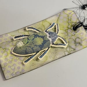 May include: A decorative tag with a watercolor background in shades of green and purple. A paper cutout of a beetle with blue and green floral patterns is affixed to the tag. The tag has a black string attached.