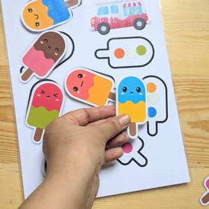 K&ouml;nnte beinhalten: Eine Hand h&auml;lt einen bunten Eis am Stiel-Aufkleber mit blauem und gelbem Design. Auf einem wei&szlig;en Blatt Papier befinden sich weitere Eis am Stiel-Aufkleber und ein rot-wei&szlig;er Van-Aufkleber. Die Aufkleber haben niedliche, cartoonartige Gesichter.