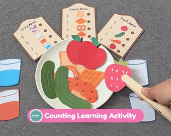 Pode incluir: Um conjunto de atividade de aprendizagem de contagem com um prato branco cheio de recortes de papel de alimentos como maçãs, pepinos, waffles e um morango, incluindo três cartões de menu de lanches com números.