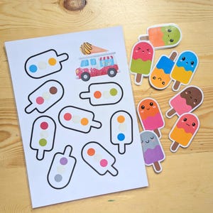 K&ouml;nnte beinhalten: Ein Bogen mit bunten Eis am Stiel-Aufklebern und einzelne Eis am Stiel-Aufkleber auf einer Holzoberfl&auml;che. Die Aufkleber zeigen verschiedene Designs, darunter l&auml;chelnde Gesichter und einen Eiswagen.