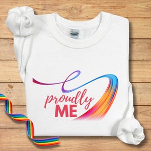 May include: White sweatshirt with the text "proudly ME" in pink and a colorful, flowing design. A rainbow ribbon is visible on the left side of the image. The sweatshirt is made of a soft material.