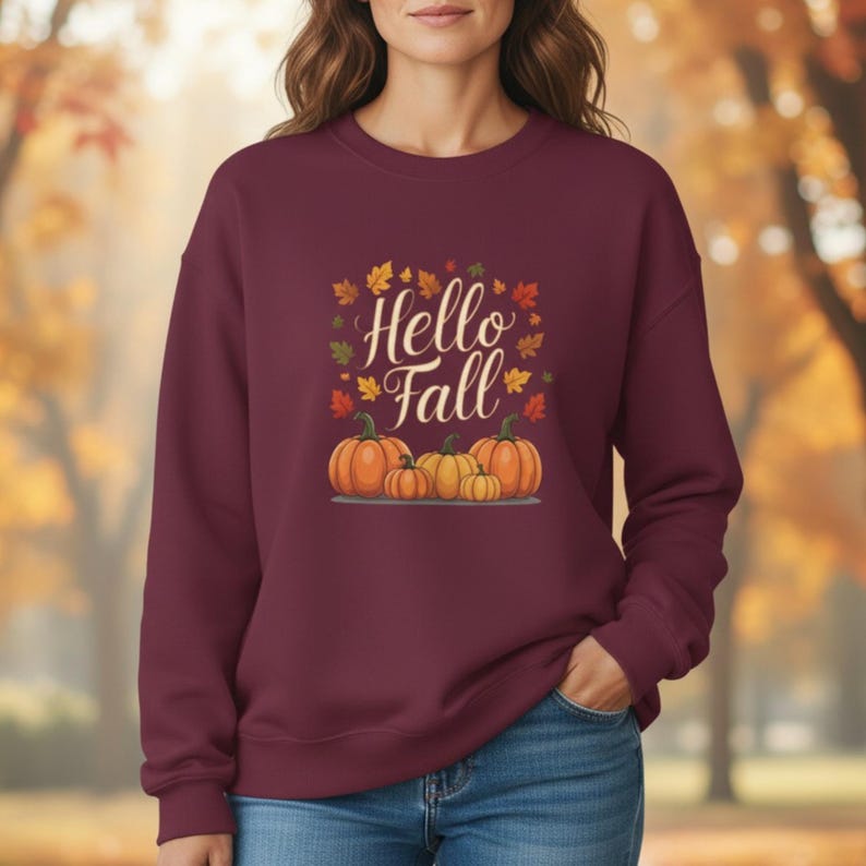 May include: A maroon crewneck sweatshirt featuring a graphic print of four orange pumpkins and scattered autumn leaves in shades of yellow, orange, and red. The text "Hello Fall" is printed in a white cursive script above the pumpkins.