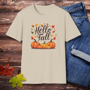 May include: A beige short-sleeve t-shirt featuring a graphic print of four orange pumpkins in a row with the text Hello Fall written in dark brown script above them, surrounded by small scattered autumn leaves in shades of red, orange, and yellow.