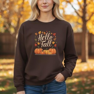May include: A dark brown crewneck sweatshirt featuring a graphic print with the text "Hello Fall" in a white script font. The design includes a cluster of four orange pumpkins of varying sizes at the base, surrounded by scattered autumn leaves.
