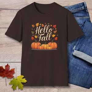 May include: A dark brown short-sleeve t-shirt featuring a graphic print of four orange pumpkins and the text Hello Fall in white cursive script, surrounded by small colorful autumn leaves in shades of red, orange, yellow, and green.