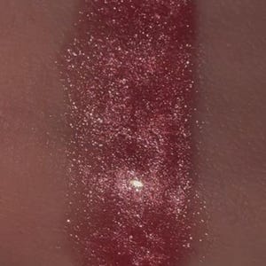 May include: Close-up of a swatch of shimmering, reddish-pink lip gloss. The gloss is applied in a rectangular shape and reflects light, showcasing its glittery texture. The background is a skin tone.