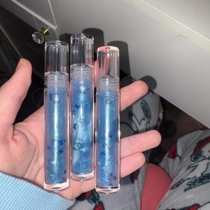 May include: Three clear lip gloss tubes filled with blue liquid and butterfly-shaped glitter. The tubes have clear caps and are held in a hand. The background is a light blue sweater and a patterned pajama.