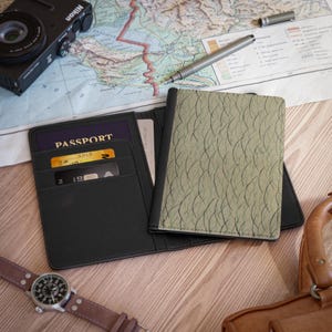 May include: Black leather passport holder with card slots, open to reveal a passport and credit cards. A green patterned passport cover is next to it. A Nikon camera, pen, and watch are also visible.