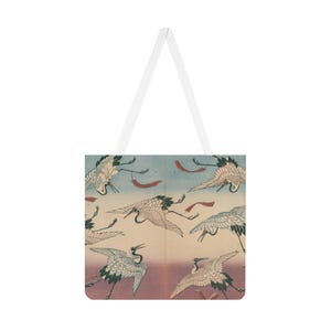 以下が含まれることがあります： 白いトートバッグ。日本の鶴のプリントが特徴で、空を飛ぶ鶴が描かれています。デザインには、青とピンクの背景に赤いアクセントが付いた様々なポーズの鶴が含まれています。白いハンドルが付いています。