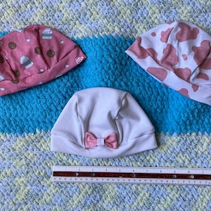 Puede incluir: Tres gorros de bebé sobre una manta colorida. Un gorro rosa tiene un estampado de galletas y cartones de leche, otro tiene un estampado de vaca y el tercero es blanco con un lazo rosa. También hay una regla presente.