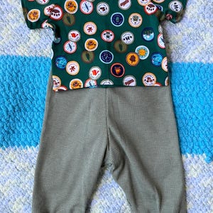Op de afbeelding: Een groen T-shirt met korte mouwen en een herhalend patroon van ronde kampeerbadges met diverse outdoor-iconen en tekst, gecombineerd met een effen olijfgroene wafelgebreide broek met boorden bij de enkels en een elastische tailleband.