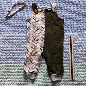 Puede incluir: Un mono de bebé con un estampado de hojas en un lado y verde oliva liso en el otro. El mono tiene puños grises y cierres de botón. También hay una diadema a juego con un estampado de hojas.