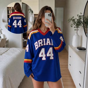 Könnte beinhalten: Ein blaues Langarmtrikot mit V-Ausschnitt. Auf der Vorderseite stehen der Text BRIAN und die Nummer 44 in Weiß. Die Ärmel haben gelbe und rote Streifen. Auf der Rückseite sind der Name GRAHAM und die Nummer 44 in Weiß mit ähnlichen Streifen.