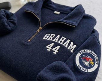 Op de afbeelding: Een marineblauwe fleece trui met een kwartrits en de naam "GRAHAM" en het nummer "44" in het wit geborduurd. Een Briar University Hawks patch staat op de mouw. De trui heeft een opvouwbare kraag en een goudkleurige rits.
