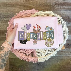 May include: A light pink sweatshirt featuring an embroidered graphic with the text Disneyland. The design includes a purple and white balloon, a pirate ship, a small elephant, a black mouse ear hat, a soft serve ice cream, and a circular icon.