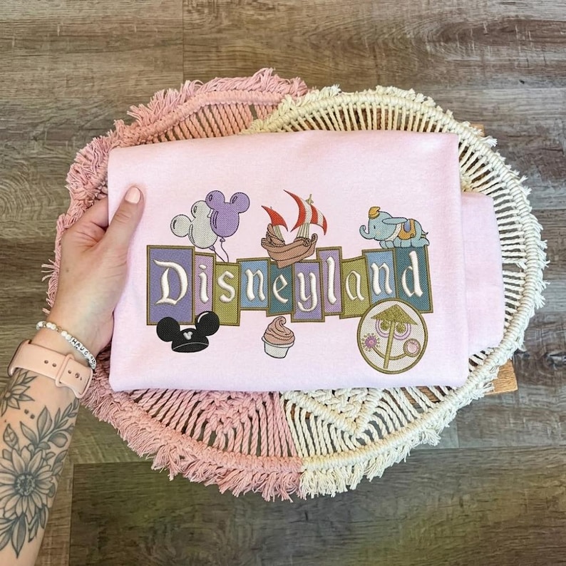 May include: A light pink sweatshirt featuring an embroidered graphic with the text Disneyland. The design includes a purple and white balloon, a pirate ship, a small elephant, a black mouse ear hat, a soft serve ice cream, and a circular icon.