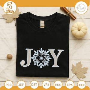 May include: Black t-shirt with the word "JOY" embroidered in light gray and white. The "O" is replaced with a snowflake design. The shirt is a Gildan brand. The image includes fall-themed decorations.