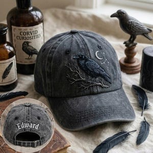 May include: A charcoal grey baseball cap featuring an embroidered black raven perched on a branch with a crescent moon and star constellation above it. The back of the cap displays the embroidered white text Edward above an adjustable metal buckle.