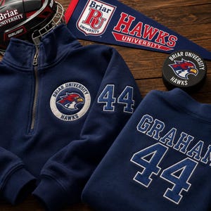 Puede incluir: Sudadera azul marino con cremallera de cuarto y sudadera de cuello redondo a juego con el texto "Briar University Hawks" y el número "44". También se ven un casco de hockey, un banderín y un disco de hockey con el mismo logotipo.
