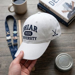Könnte beinhalten: Eine cremefarbene Baseballkappe mit dem Text "Briar 2026 University" in Marineblau. Die Kappe hat auch eine kleine Hockey-Schläger-Grafik. Der Hut ist ein klassisches Sechs-Panel-Design.