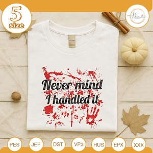 May include: White t-shirt with the text "Never mind I handled it" in black script, surrounded by red splatter and a handprint design. The shirt has a crew neck and is displayed on a wooden surface with fall-themed decorations.