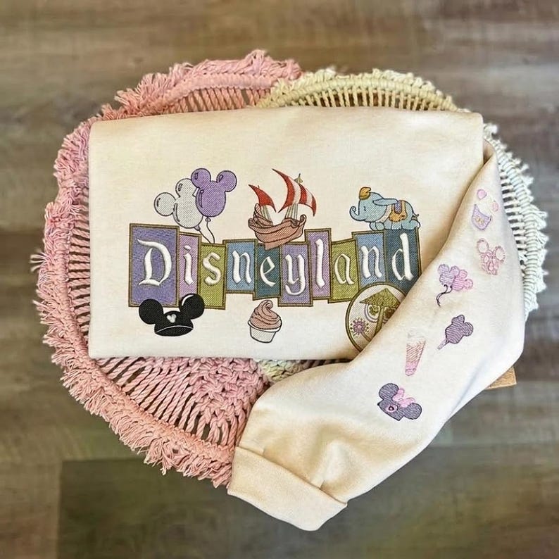 May include: A cream-colored sweatshirt featuring an embroidered Disneyland logo in block letters. The design includes colorful icons like balloons, a ship, Dumbo, a cupcake, and a carousel. The sleeve displays smaller embroidered Disney-themed icons.