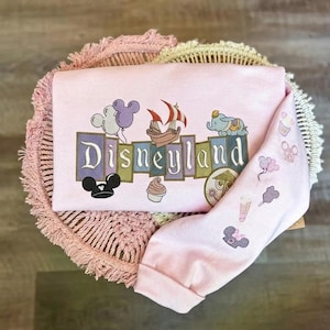 May include: A light pink long-sleeve sweatshirt featuring an embroidered Disneyland logo in block letters. The design includes icons of a ship, Dumbo, Mickey ears, and a cupcake. The sleeve features smaller embroidered icons of balloons and treats.