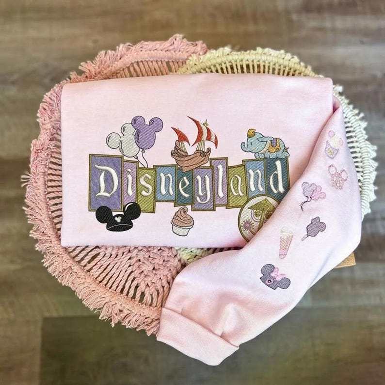 May include: A light pink long-sleeve sweatshirt featuring an embroidered Disneyland logo in block letters. The design includes icons of a ship, Dumbo, Mickey ears, and a cupcake. The sleeve features smaller embroidered icons of balloons and treats.