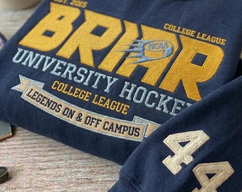 Op de afbeelding: Marineblauw sweatshirt met goudkleurige en witte geborduurde tekst. De tekst luidt "BRRR UNIVERSITY HOCKEY COLLEGE LEAGUE LEGENDS ON & OFF CAMPUS" met het nummer "44" op de mouw. De sweatshirt heeft een vintage college-stijl.