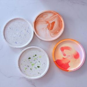 May include: Four round decorative trays in shades of white, orange, and pink. Two trays have a speckled design, while the other two feature swirled patterns. The trays are made of a solid material and are suitable for holding small items.